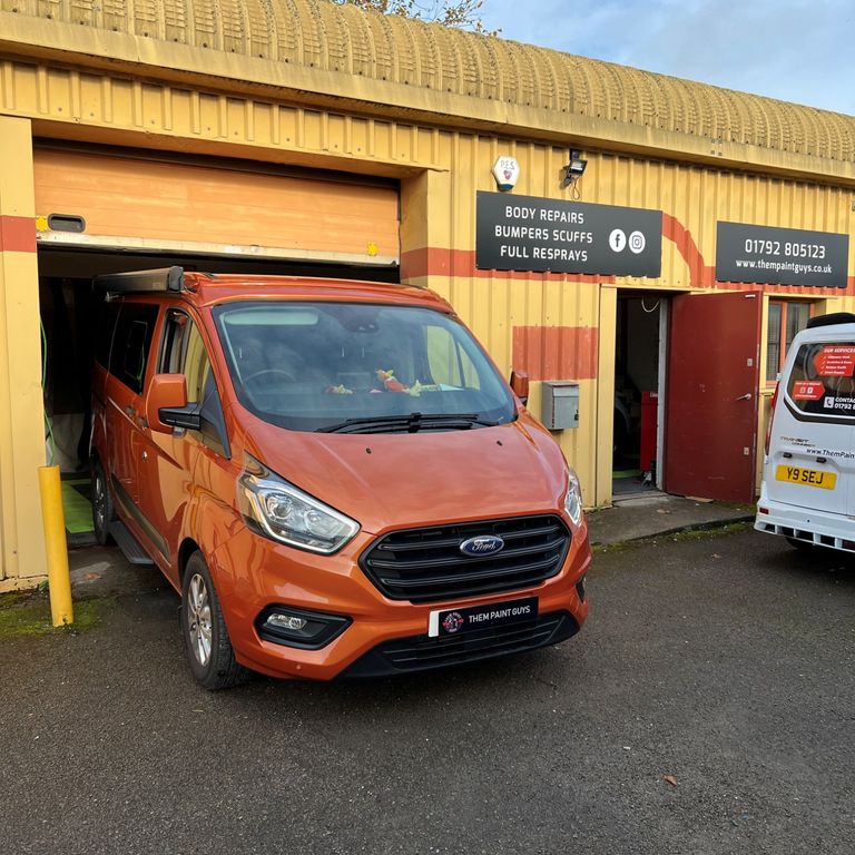 A signaged van is parked outside Them Paint Guys garage, with a second orange van parked partially inside the shutter after being repaired